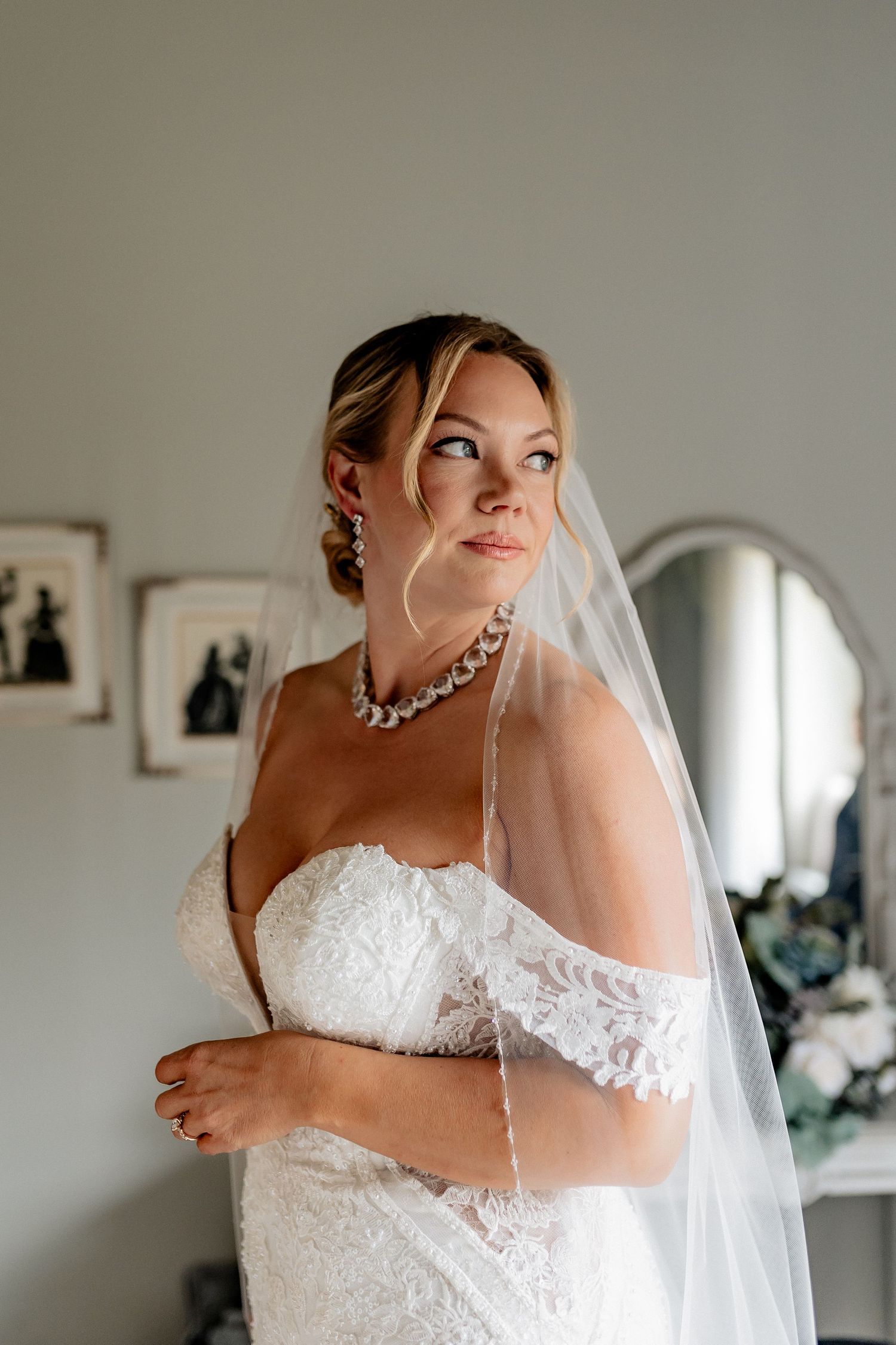 A bride in an elegant white lace wedding dress with a veil and statement necklace poses thoughtfully in a dressing room. Hawthorne Estates