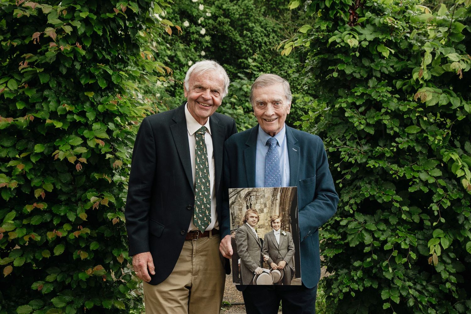 Outdoor portrait at The Oaksmere in Suffolk of two men holding a photo of themselves from the wedding 50 years earlier.