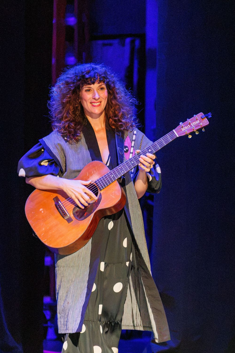 A curly-haired woman smiles while playing an acoustic guitar on stage, wearing a polka dot dress and gray cardigan.