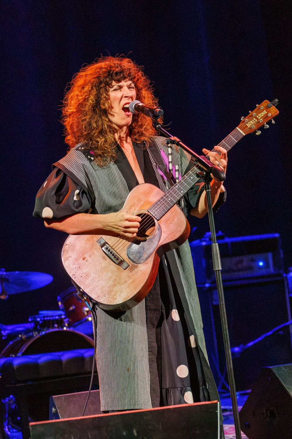 Red-haired woman performing on stage, playing acoustic guitar and singing into microphone, wearing polka dot dress.