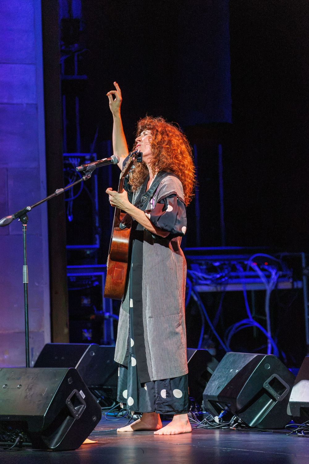 Curly-haired female guitarist performs barefoot on stage, raising one hand while singing into a microphone at a live concert.