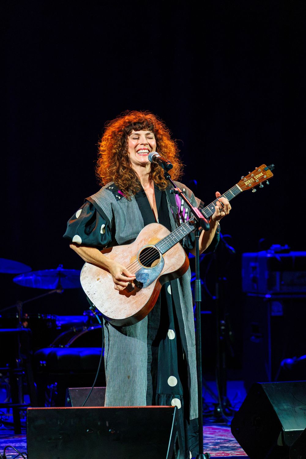 Curly-haired woman smiling while playing acoustic guitar on stage in a dark floral kimono during a live concert.