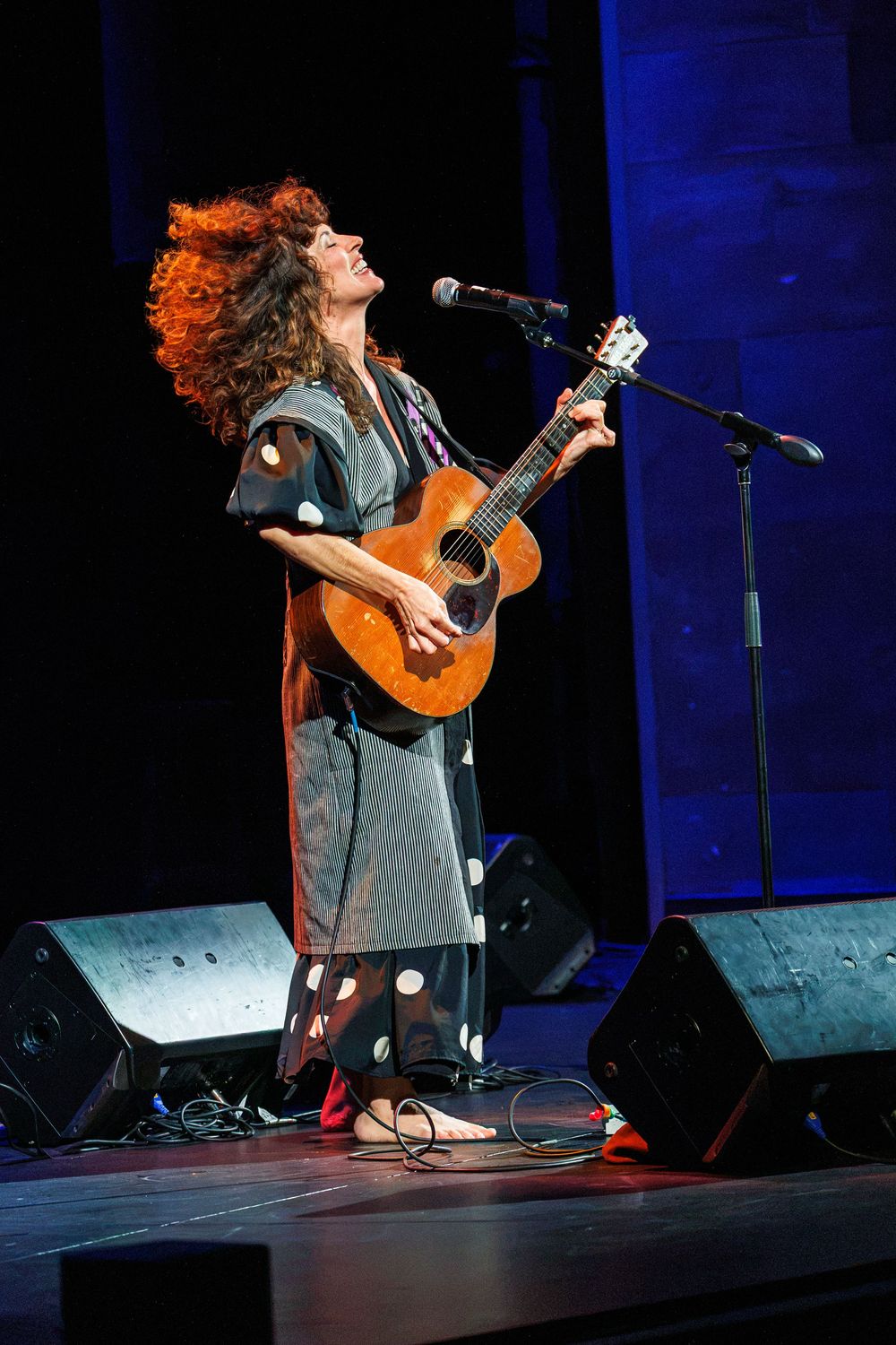 Curly-haired woman playing acoustic guitar and singing on stage during a live concert performance.