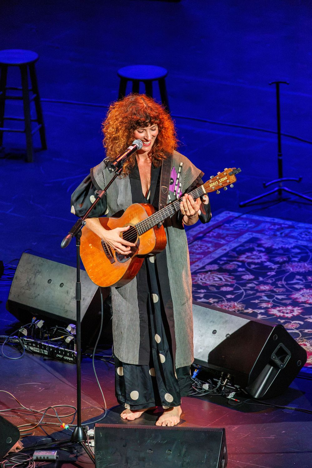 Red-haired woman playing acoustic guitar barefoot on stage at a live concert with blue stage lighting.