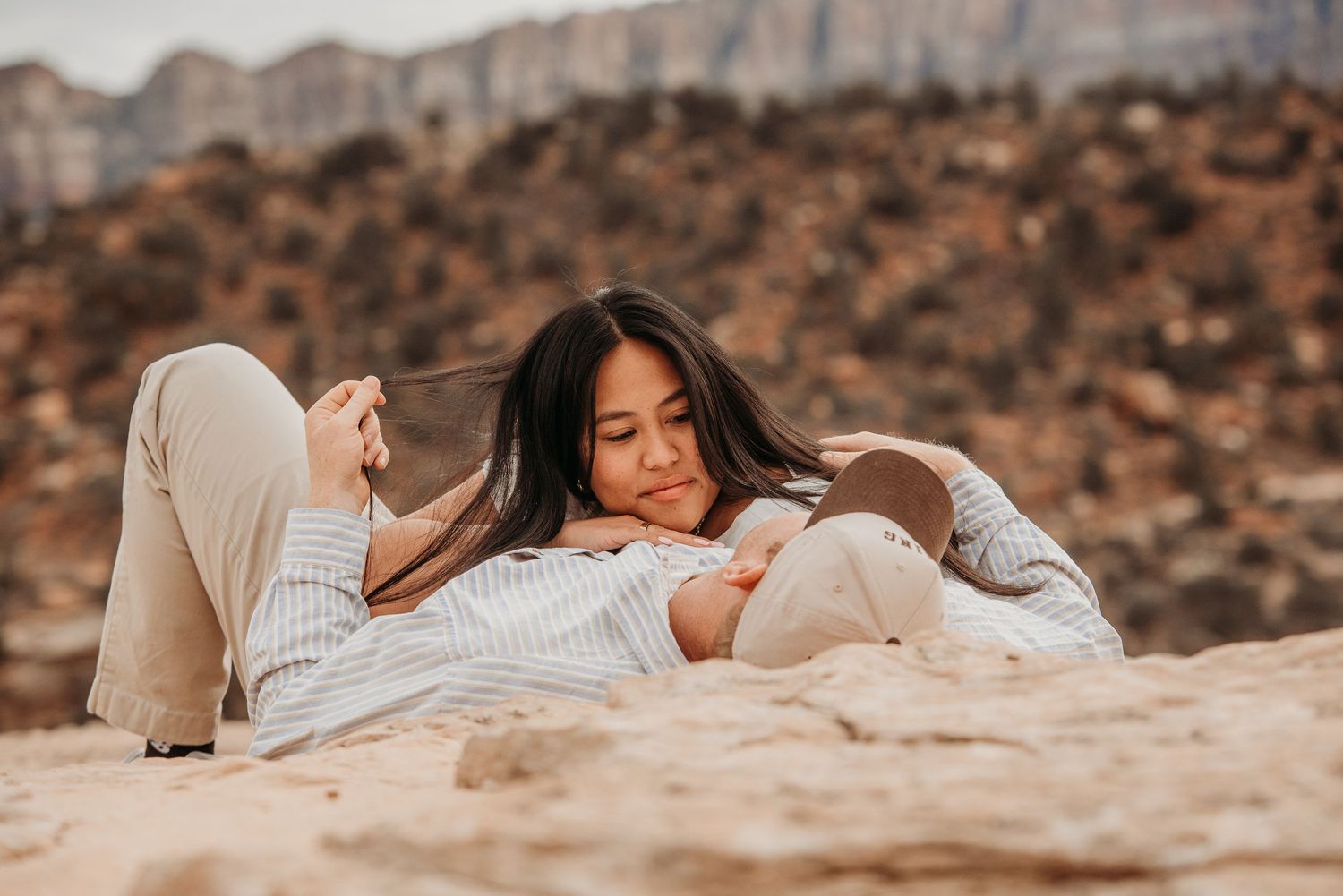 Woman lying on sandy rock in desert landscape, wearing striped shirt, relaxing outdoors with mountains in background.