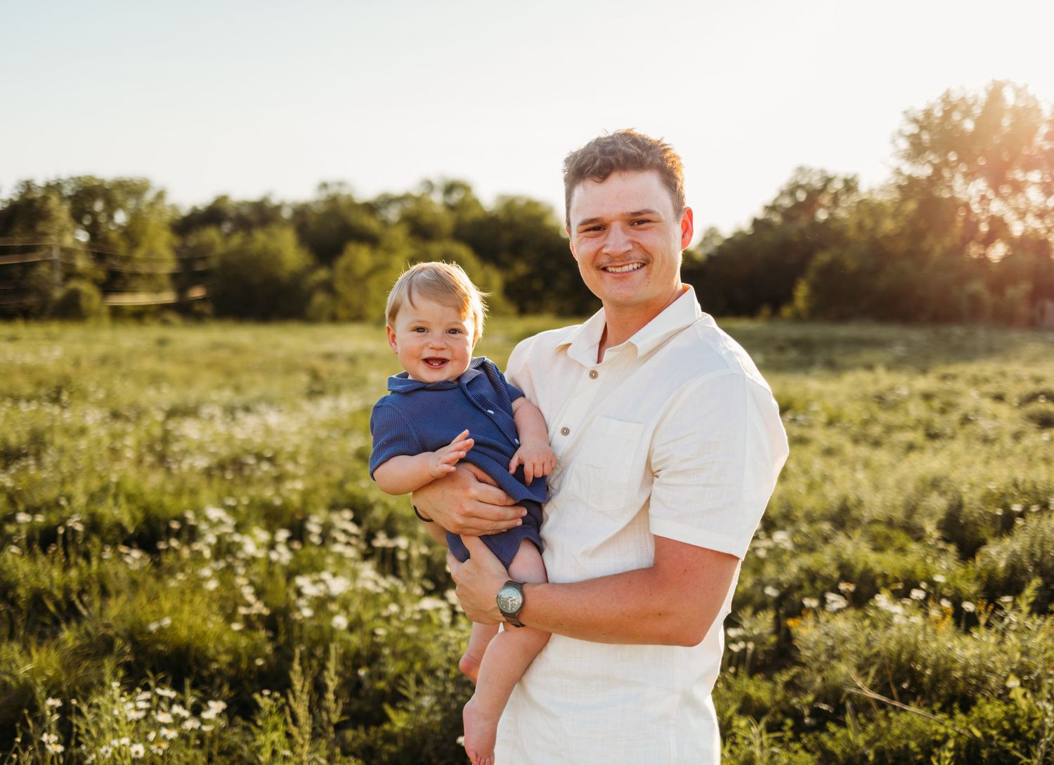 A smiling man holds a happy baby in a sunny green field, perfect for family portrait photography outdoors.
