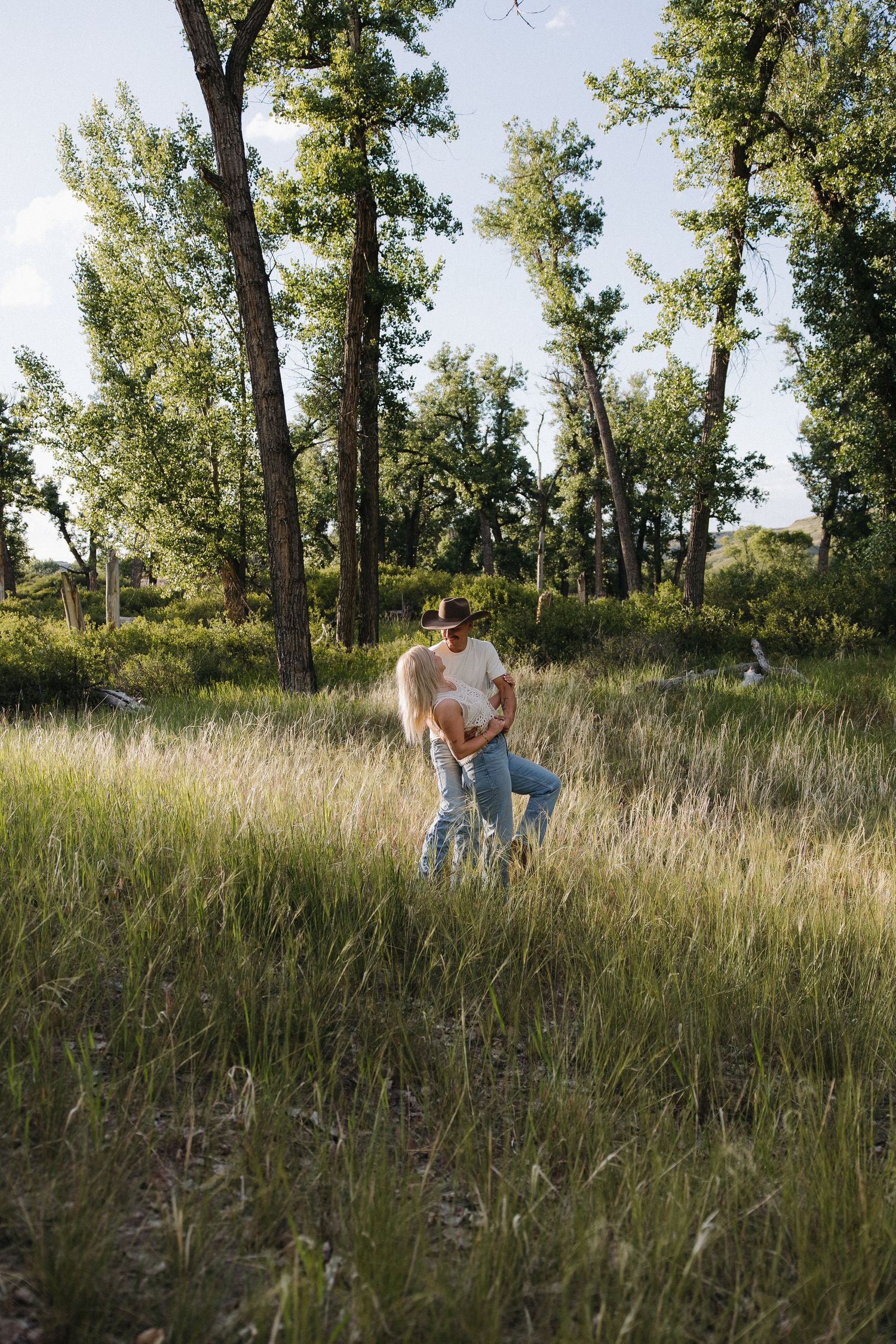 Couple embracing in sunlit tall grass field surrounded by green trees on a sunny day.