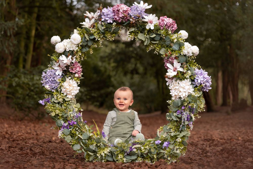 Baby sitting inside a floral wreath arch with purple and white flowers outdoors.