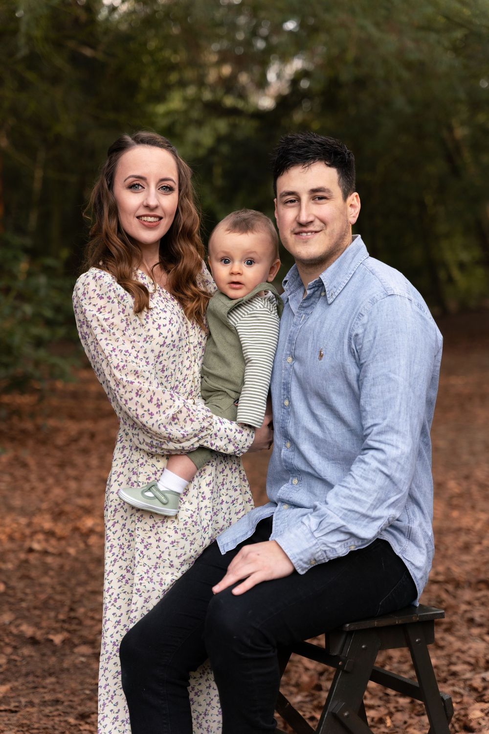 Young family portrait with mother, father, and baby posed outdoors among autumn leaves in a woodland setting.