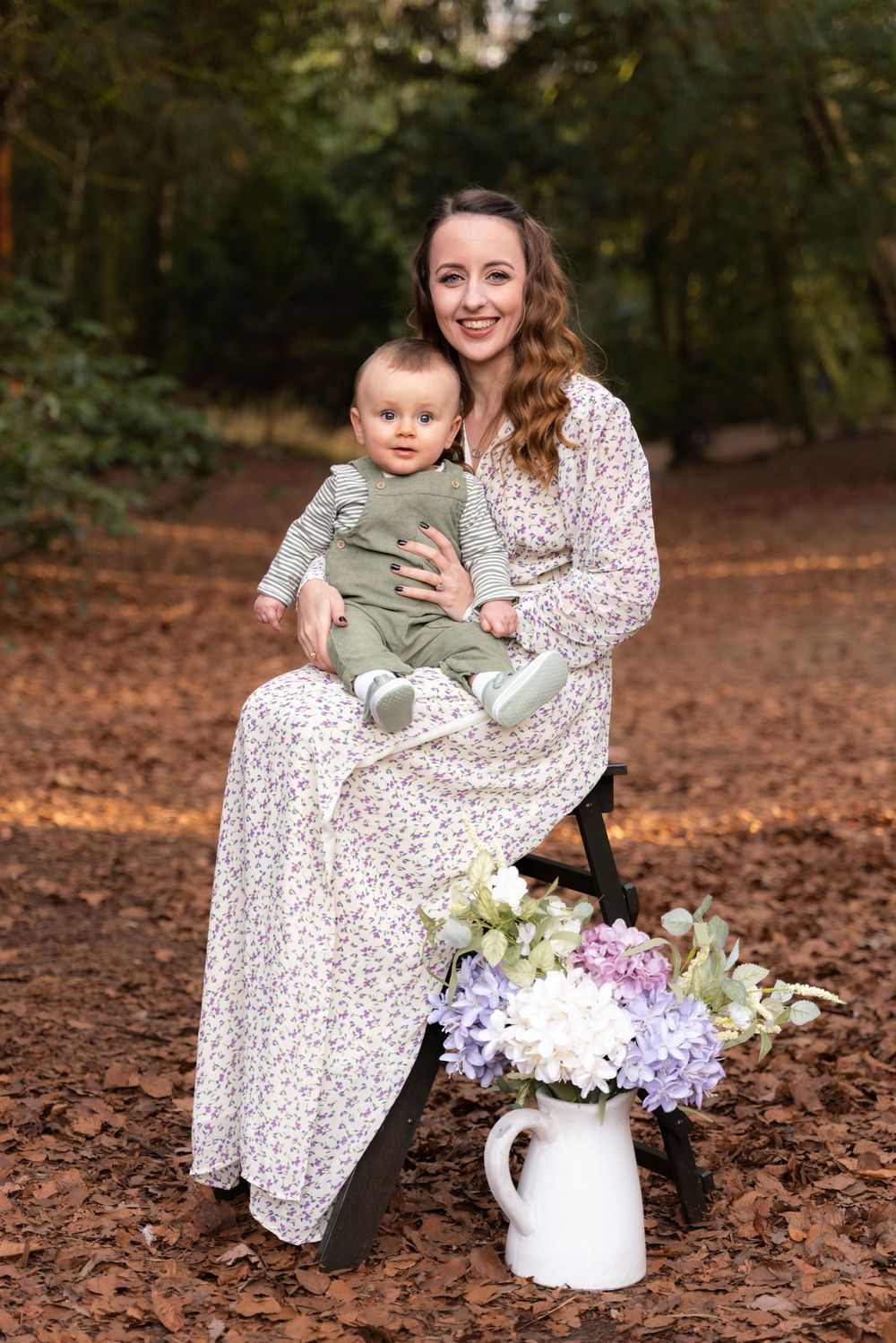 Smiling woman in floral dress holds baby in green outfit, seated outdoors among autumn leaves with white flower jug.