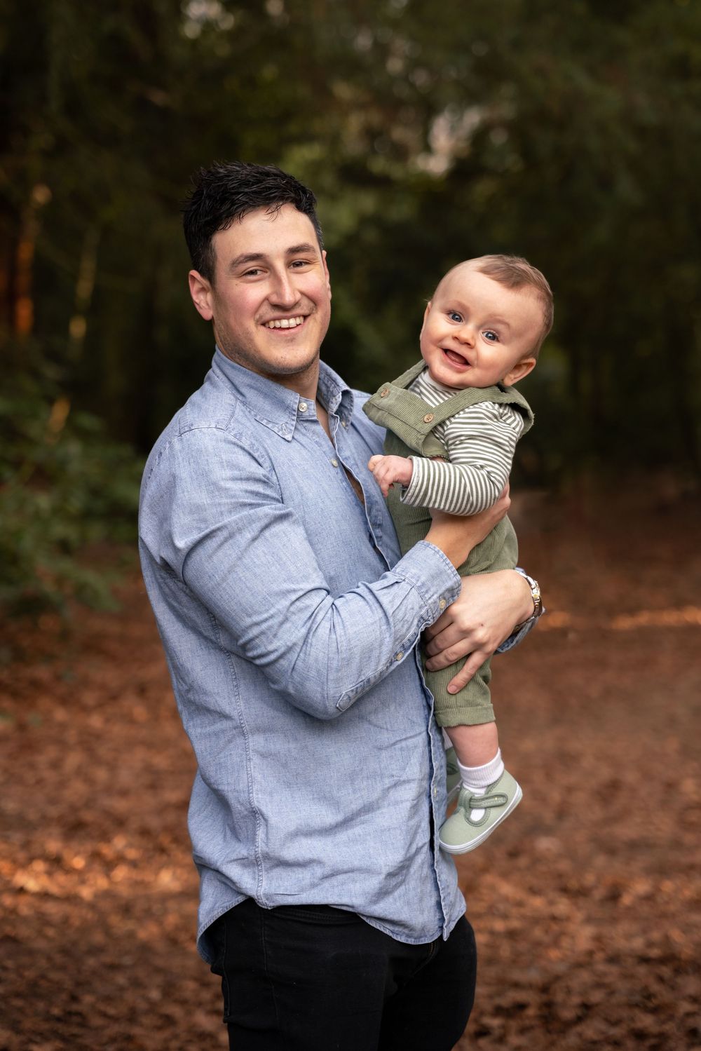 Smiling young man in blue shirt holding a happy baby in green outfit on a forest path.