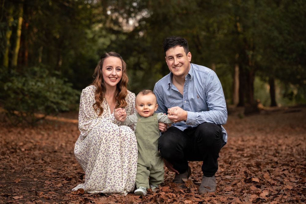 A young family with a baby boy poses outdoors on autumn leaves in a woodland setting for a professional photo.