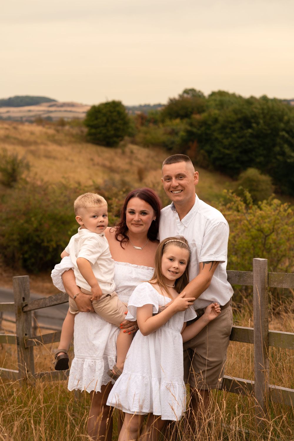 Family of four poses outdoors near a wooden fence in golden fields, parents and two young children wearing white outfits.