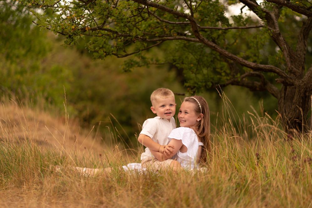 Two young children in white outfits sitting together in golden grass beneath a tree outdoors.