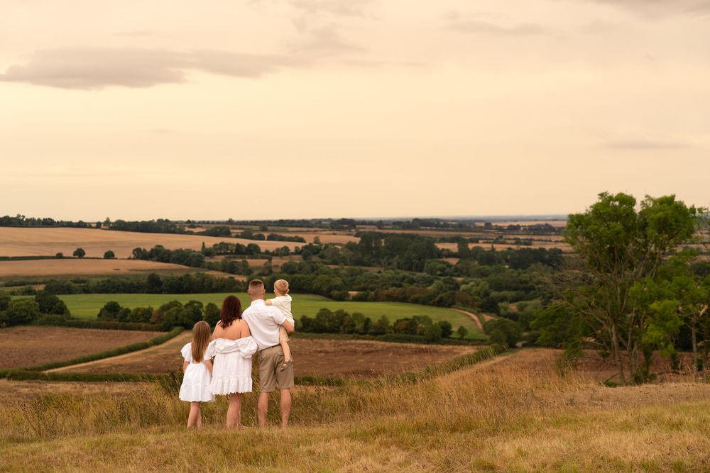 Family of three standing on a hill overlooking a scenic countryside landscape at sunset.