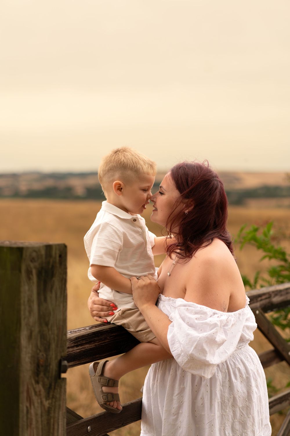 A mother in a white off-shoulder dress shares a tender moment with her young son seated on a wooden fence in a rural field.