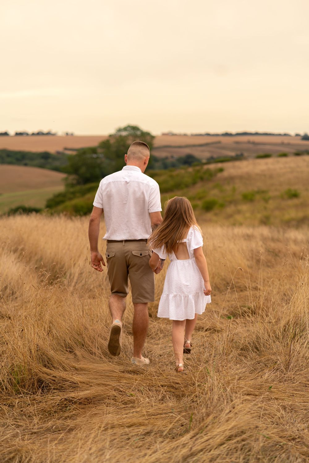 A man and young girl in white outfits hold hands walking through a golden field at dusk.