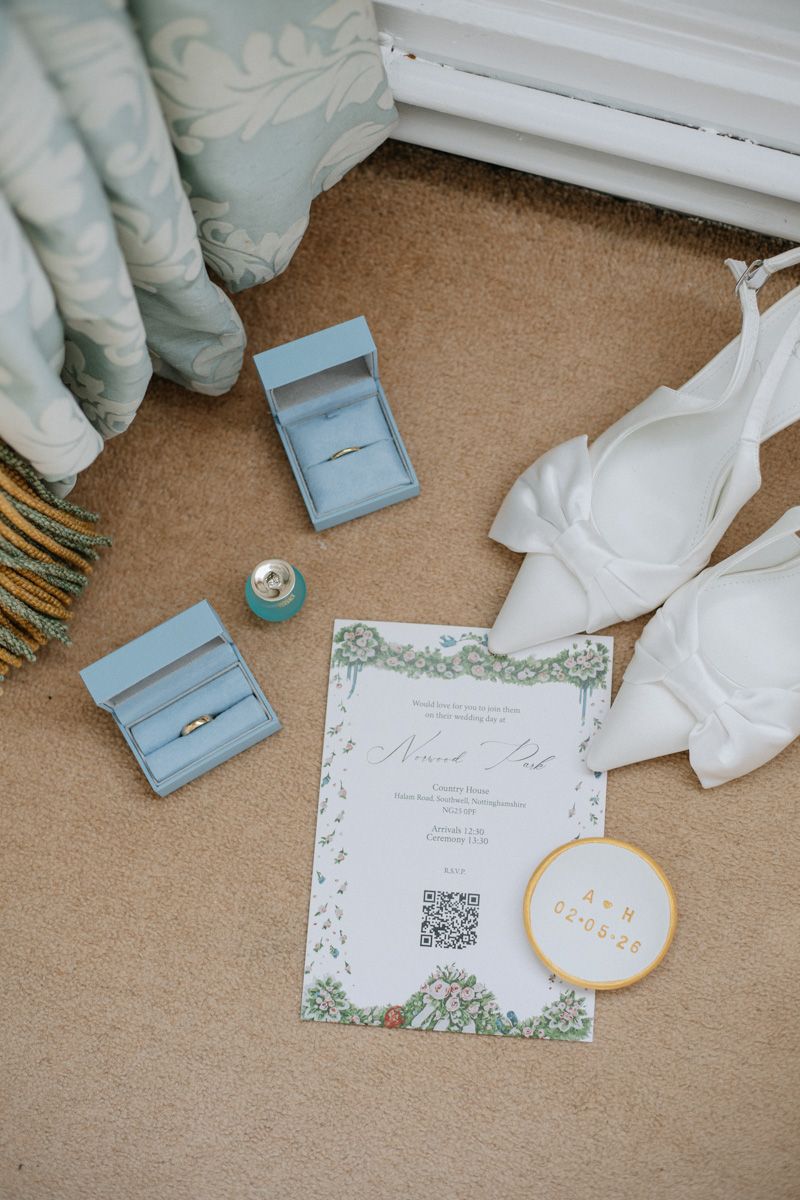 Wedding flat lay with rings in blue boxes, invitation, white heels, and gold details on carpet.