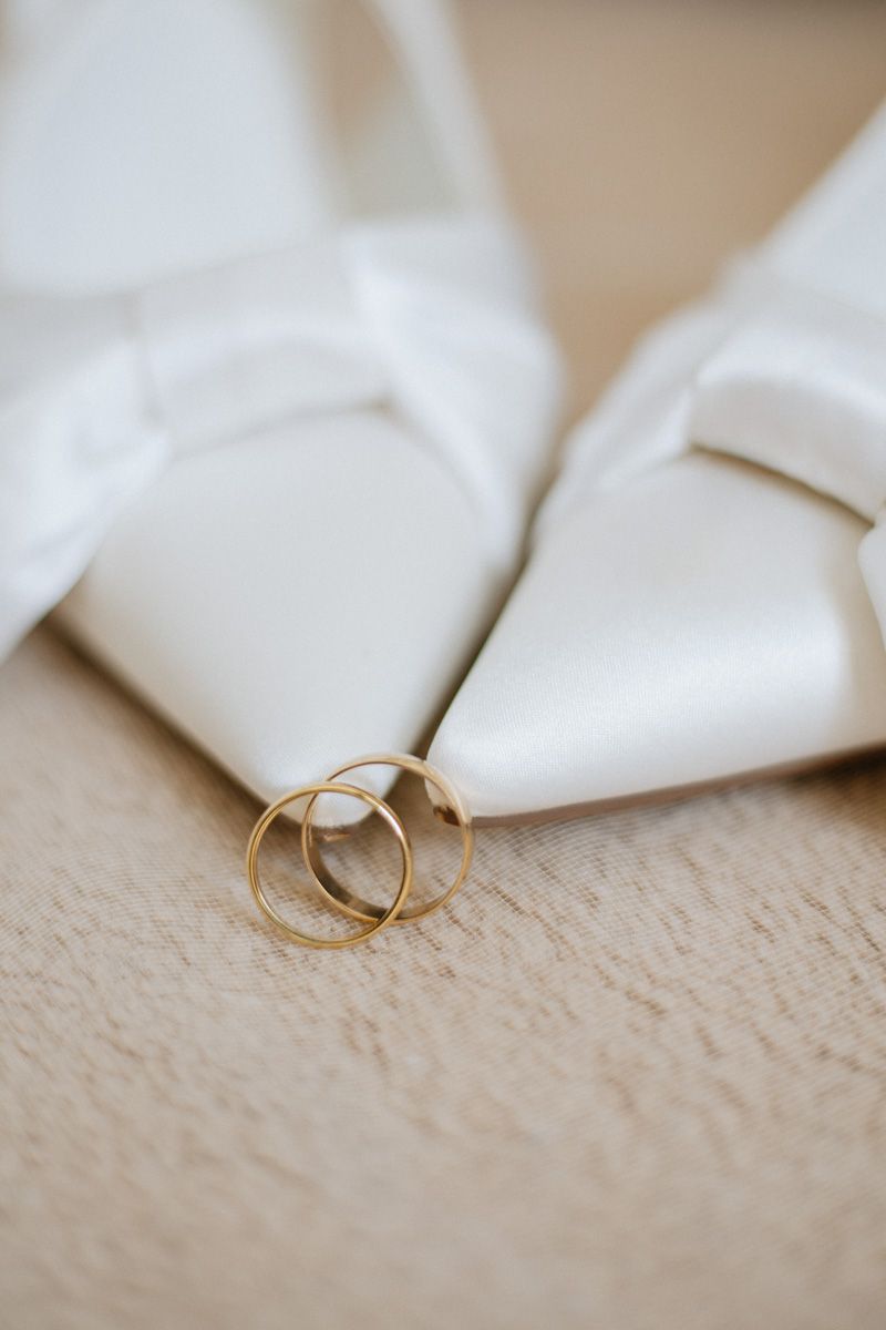 Two gold wedding bands resting against white satin bridal heels with bow detail on a neutral surface.
