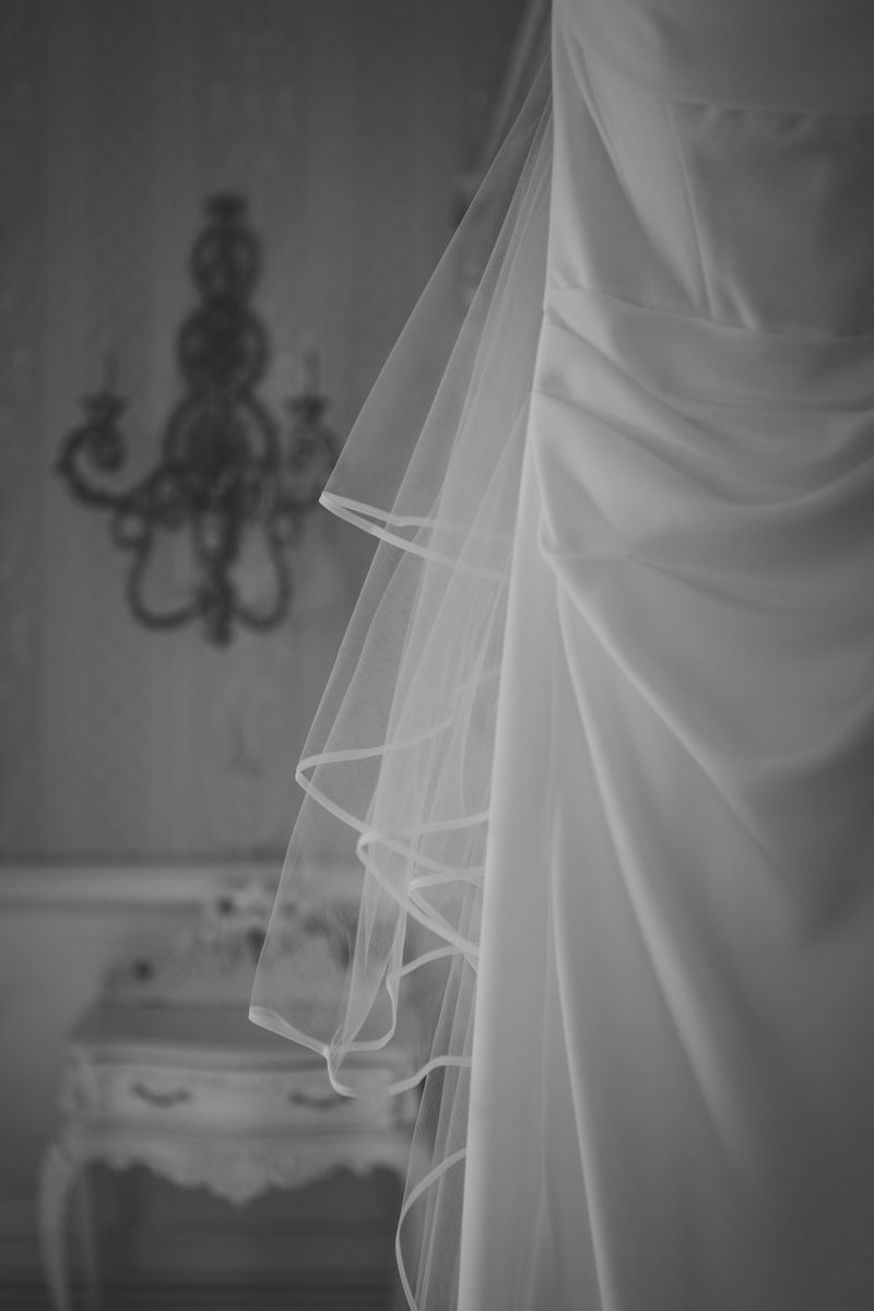 Black and white photo of a bridal veil and wedding dress hanging near an ornate candelabra wall sconce.