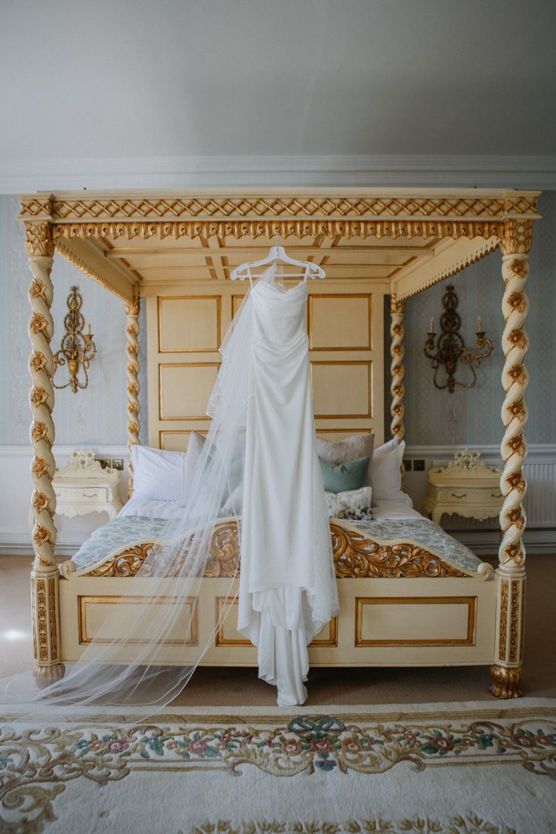 Elegant white wedding dress hanging on a ornate gold four-poster bed in a luxurious bridal suite.