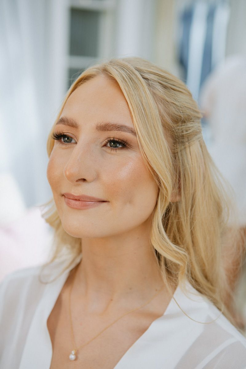 Blonde woman with soft makeup, pearl necklace, and half-up hairstyle smiling gracefully in white attire.
