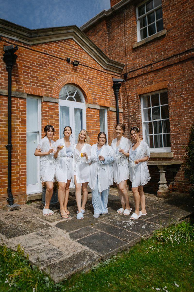 Six bridesmaids in white robes holding champagne glasses outside a red brick manor house on a wedding day.