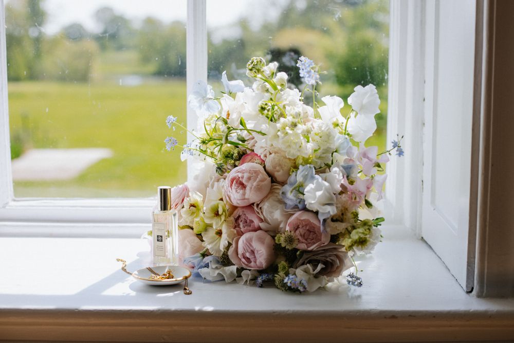 Romantic bridal bouquet of blush peonies and white flowers on a windowsill with perfume bottles.