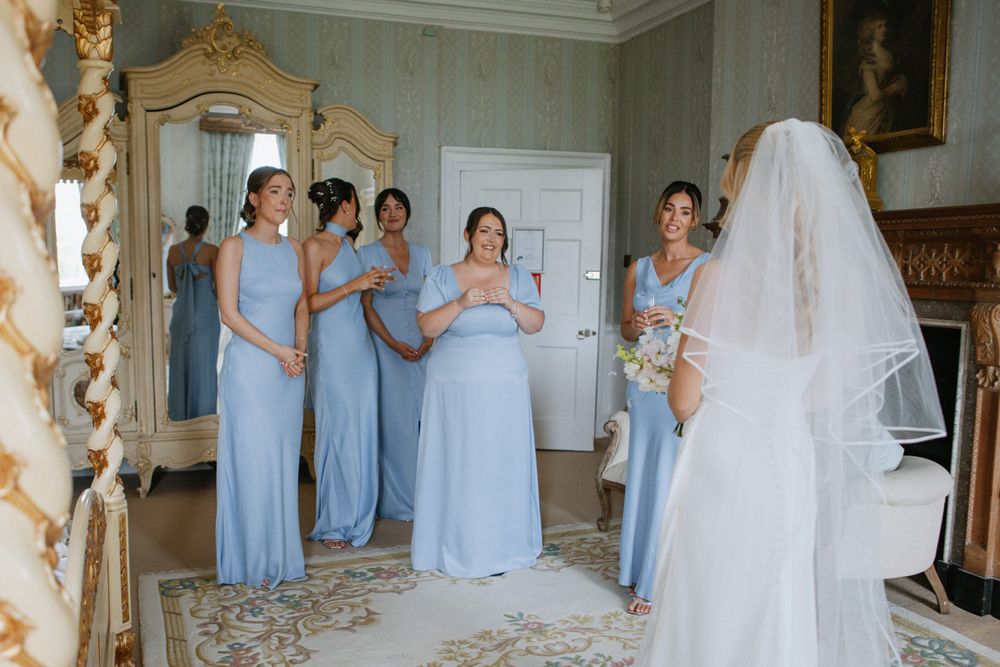Bridesmaids in light blue gowns react with joy as bride in white gown and veil reveals her look in elegant room.