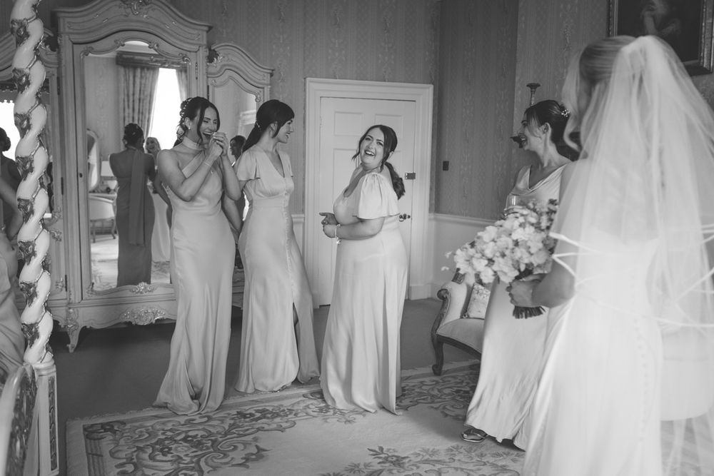 Bridesmaids in elegant gowns react emotionally as bride in white dress and veil reveals her look in a classic wedding venue.