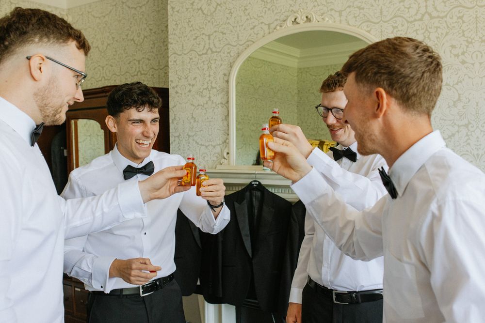 Four groomsmen in white shirts and bow ties toasting with amber bottles during wedding preparations.