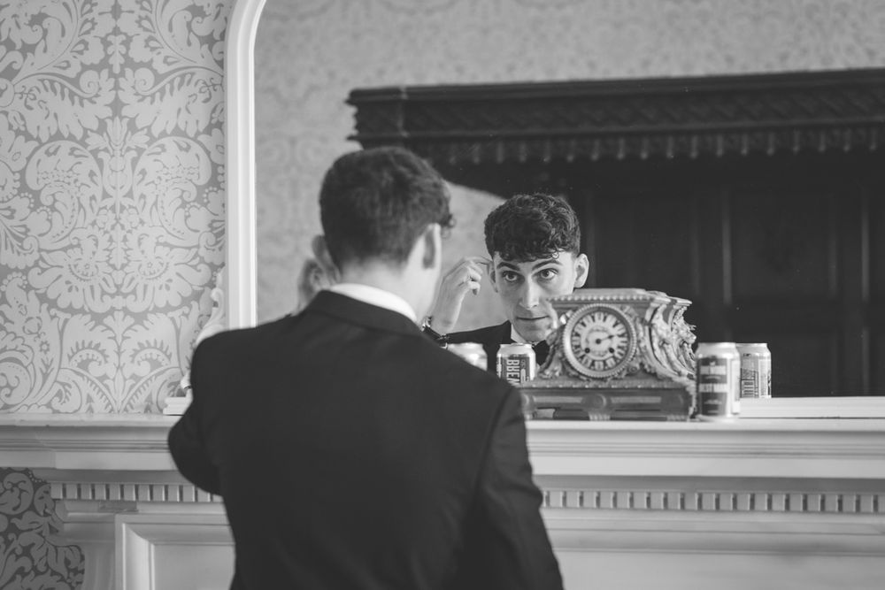 Groom adjusting his hair in an ornate mirror above a fireplace mantel in a classic interior.