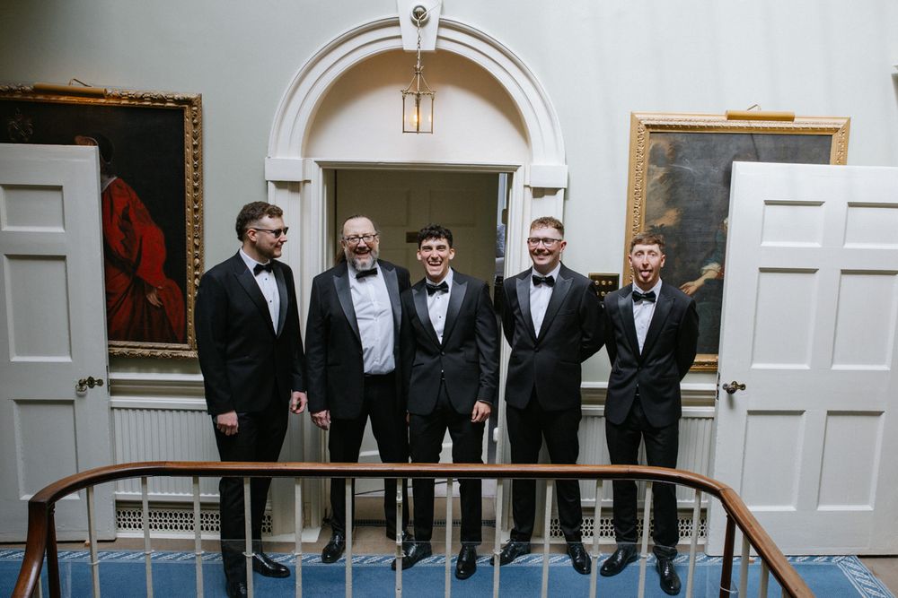 Five men in black tuxedos and bow ties posing together on a staircase landing in an elegant venue.