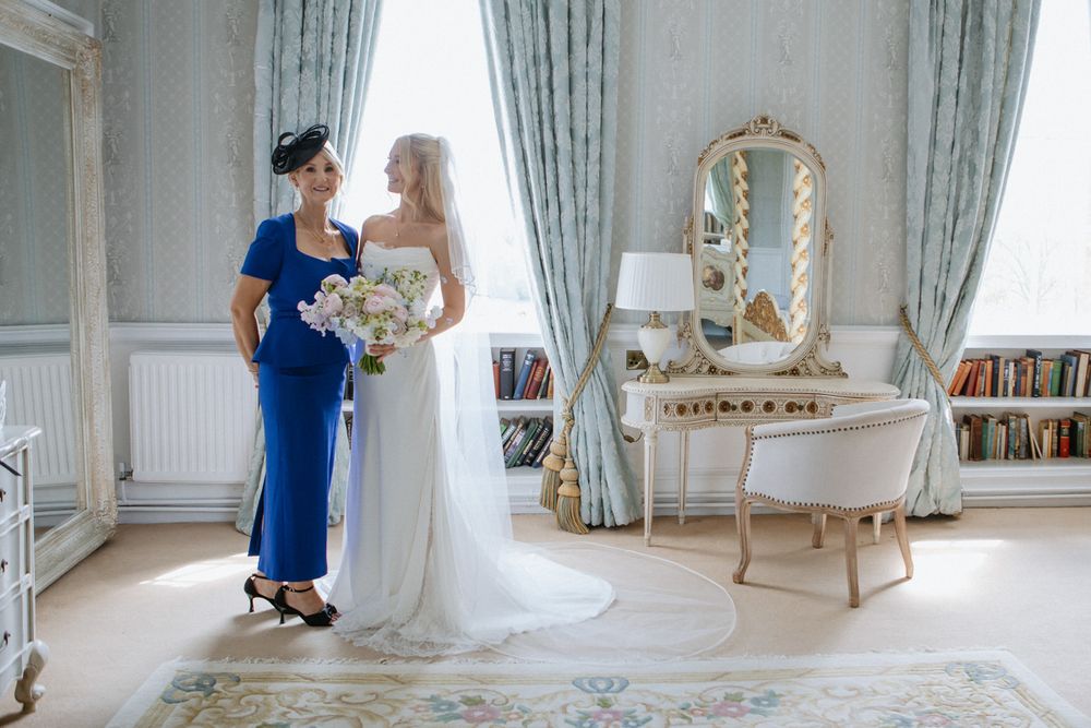 Bride in white gown and woman in blue dress holding bouquet in elegant room with mirror and drapes.