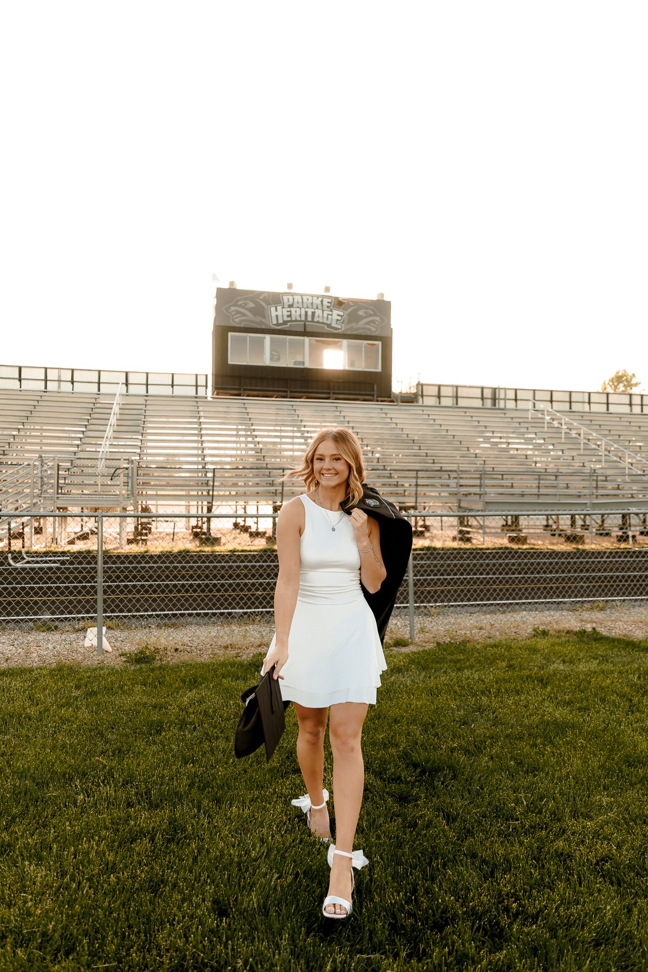 Smiling young woman in white dress holding graduation cap at Heritage high school football stadium.