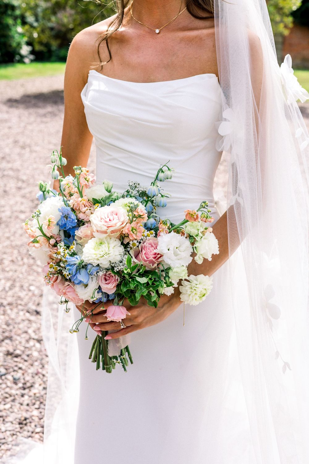 Bride in strapless white wedding gown holding a lush bouquet of pink, blue, and white flowers with greenery.