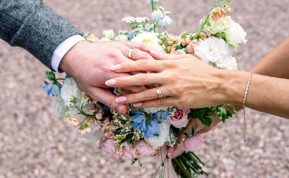 Bride and groom hands resting on wedding bouquet of pink and blue flowers, showing wedding rings.