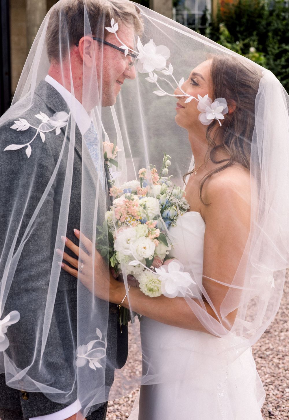 Bride and groom share romantic moment under floral embroidered veil, bride holding pastel bouquet.