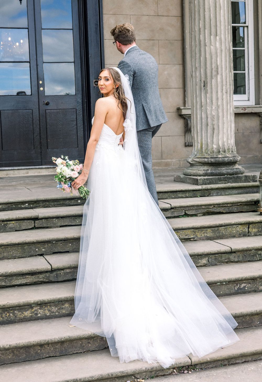 Bride in a white tulle gown with bouquet and groom in grey suit on stone steps of an elegant venue.