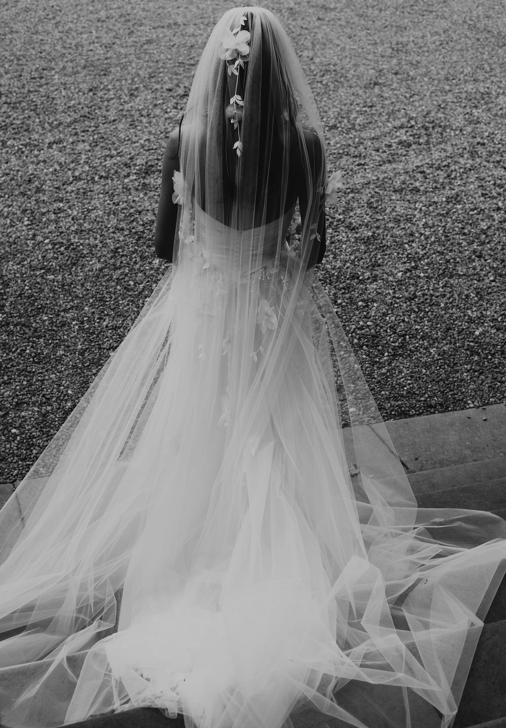 Black and white photo of a bride in a flowing wedding gown and long veil with floral accents, viewed from behind.
