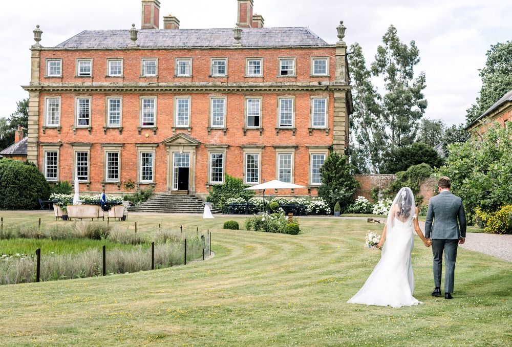 Bride and groom walk hand-in-hand toward a grand red-brick Georgian country house wedding venue surrounded by manicured lawns.