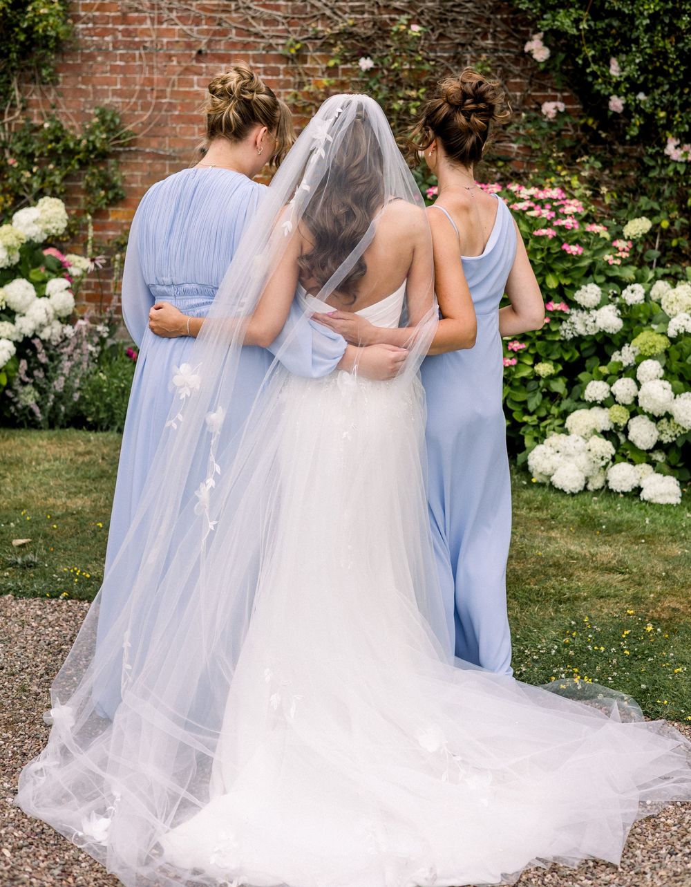 Bride in white gown and veil flanked by two bridesmaids in blue dresses, standing in a garden with white flowers.