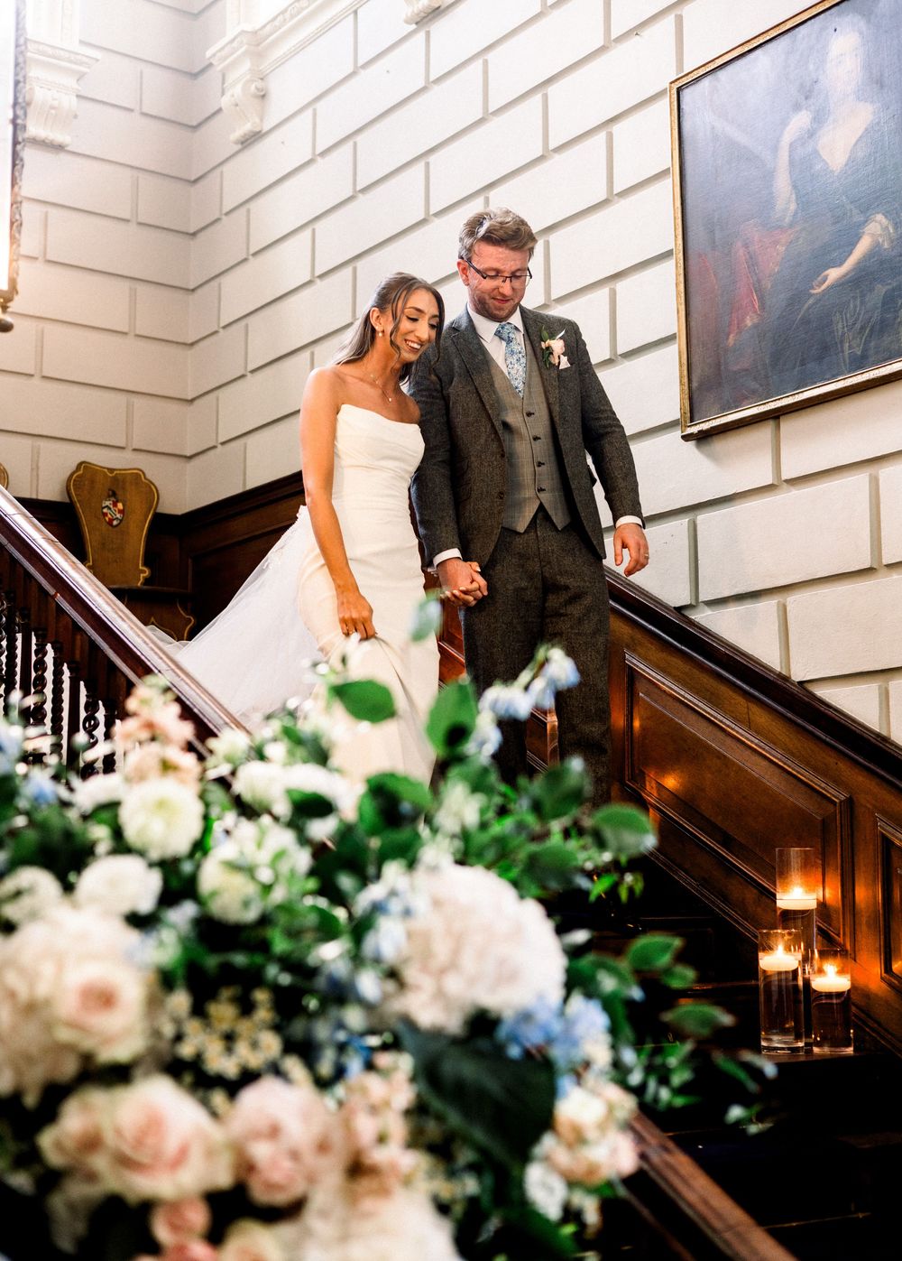 Bride and groom descend grand staircase adorned with candles and white floral arrangements at elegant wedding venue.