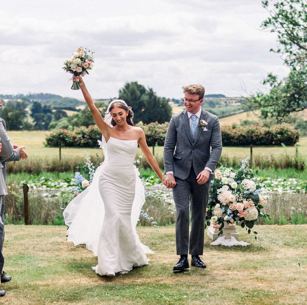 Joyful bride raises bouquet while holding groom's hand after outdoor wedding ceremony on lush green countryside lawn.