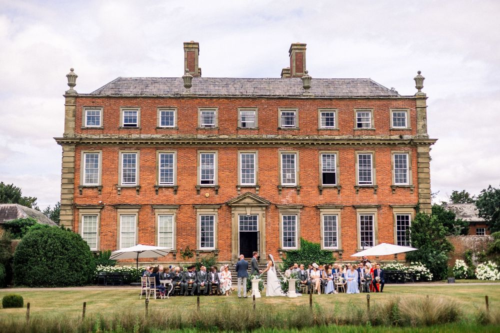 Wedding ceremony on lawn of grand Georgian red brick country house mansion with guests gathered outside.