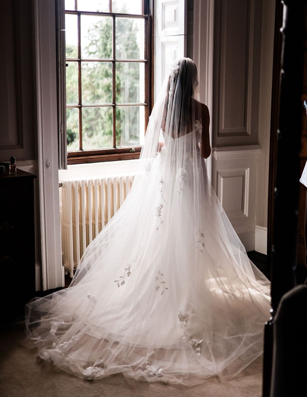Bride in a flowing white wedding gown with floral veil gazing out a large window in an elegant room.