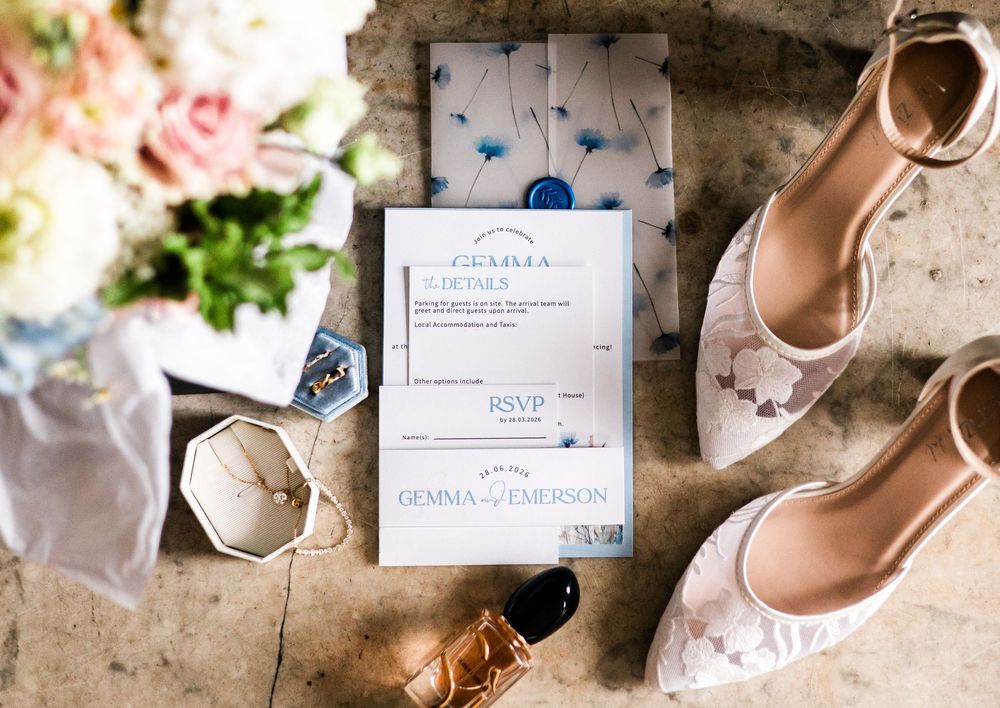 Elegant wedding flat lay featuring blue floral invitations, RSVP cards, lace heels, jewelry, and pink roses on marble.