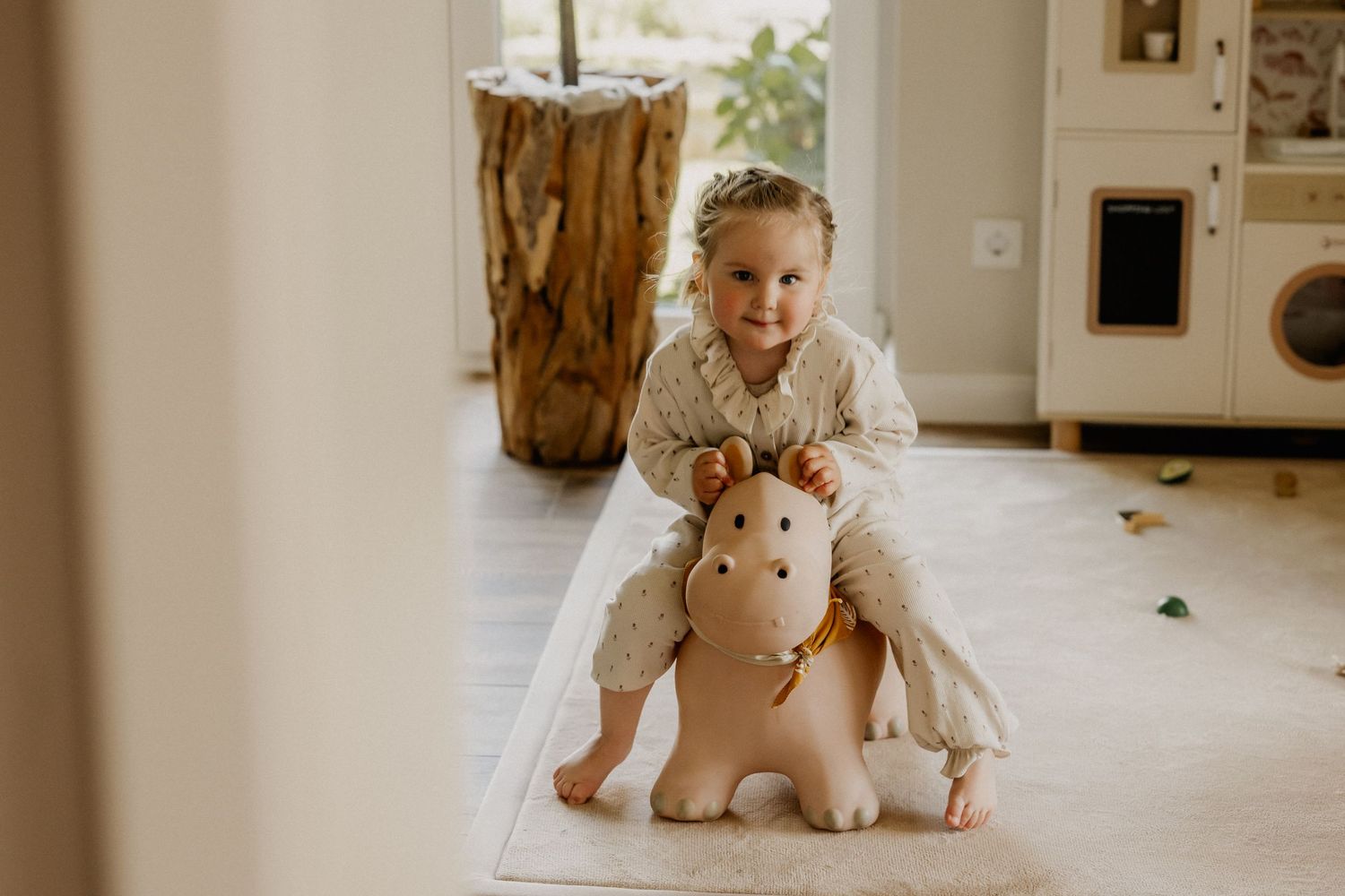 Young girl with braids smiling while riding a beige toy horse bouncer in a bright, modern playroom.