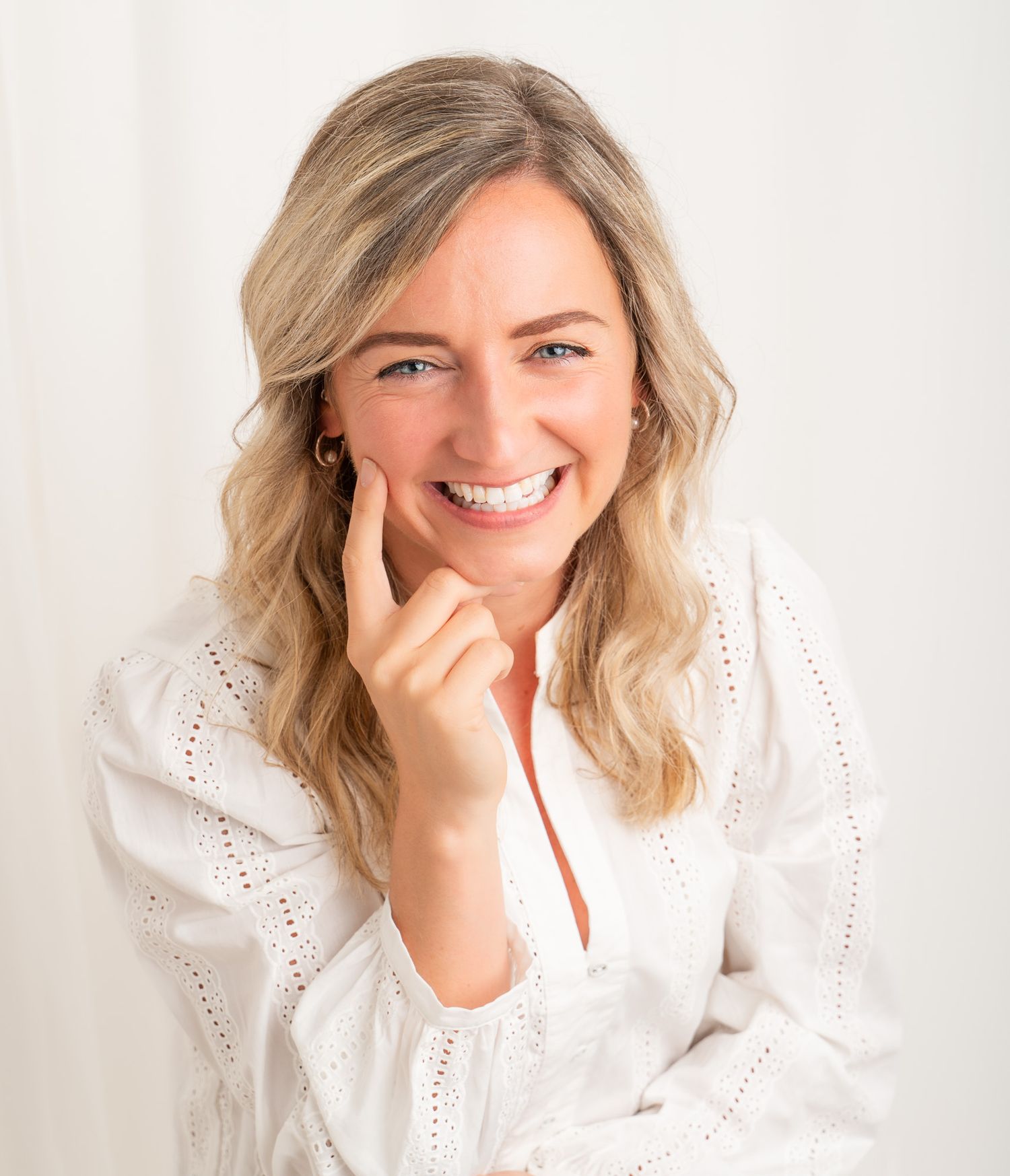 Smiling blonde woman in white eyelet blouse posing with hand near chin against light background.