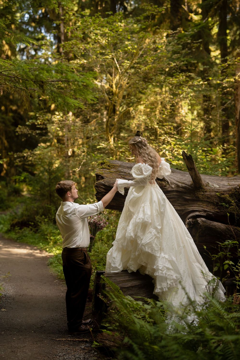 Groom helps bride in white lace gown climb a fallen log on a forest path surrounded by lush greenery.